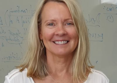 Smiling person with shoulder-length blonde hair, wearing white blouse and necklace.