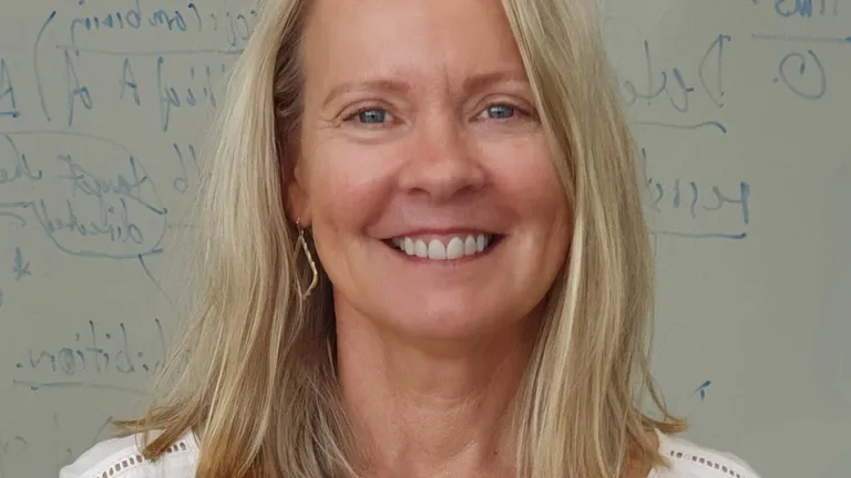 Smiling person with shoulder-length blonde hair, wearing white blouse and necklace.
