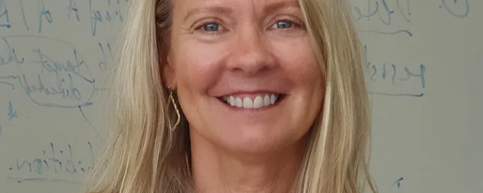 Smiling person with shoulder-length blonde hair, wearing white blouse and necklace.