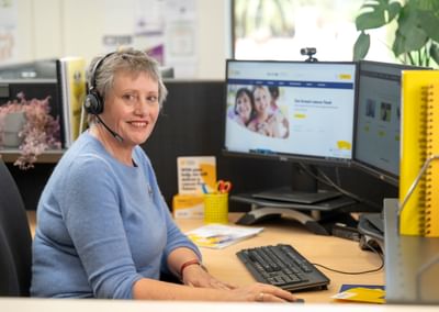 Person wearing a blue sweater and headset sits at a desk with two monitors in a bright office.