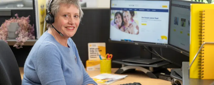 Person wearing a blue sweater and headset sits at a desk with two monitors in a bright office.