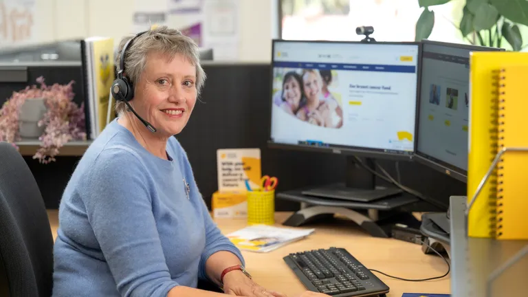 Person wearing a blue sweater and headset sits at a desk with two monitors in a bright office.