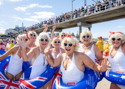Seniors in white swimsuits with blonde wigs and sunglasses, Aussie flag inflatables on the beach.