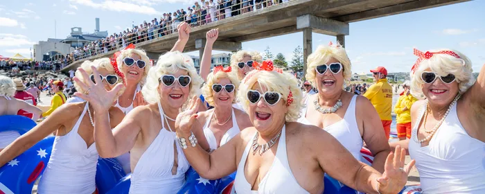 Seniors in white swimsuits with blonde wigs and sunglasses, Aussie flag inflatables on the beach.