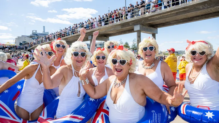 Seniors in white swimsuits with blonde wigs and sunglasses, Aussie flag inflatables on the beach.