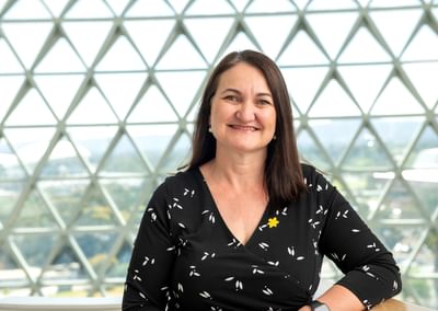 Smiling person with dark hair in a black wrap top with white leaves, a yellow pin, in a glass dome.