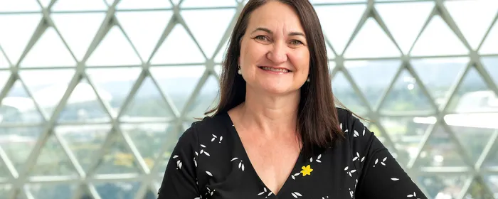 Smiling person with dark hair in a black wrap top with white leaves, a yellow pin, in a glass dome.