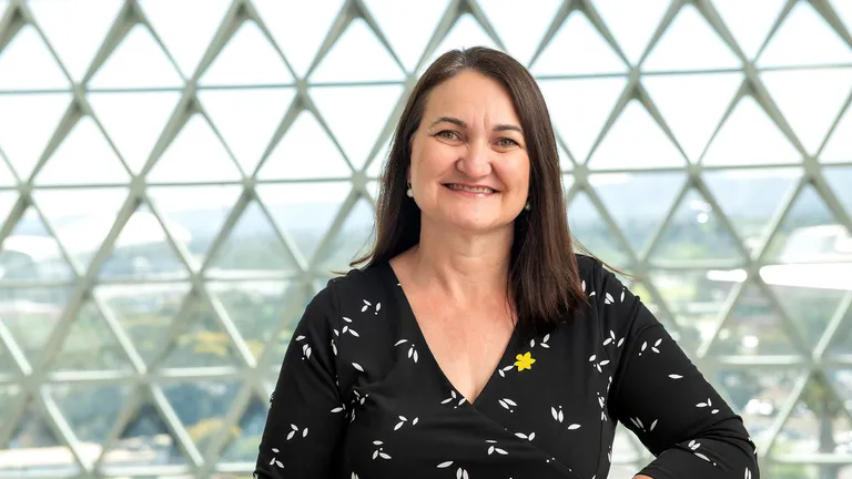 Smiling person with dark hair in a black wrap top with white leaves, a yellow pin, in a glass dome.
