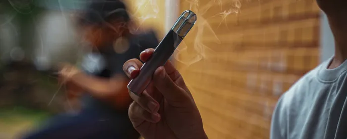 A person holds a vape pen with wispy smoke, blurred outdoor scene and brick wall.