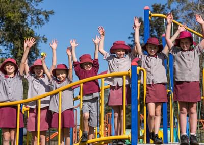 Group of children in maroon hats and checkered uniforms on a yellow-rail playground, arms raised.