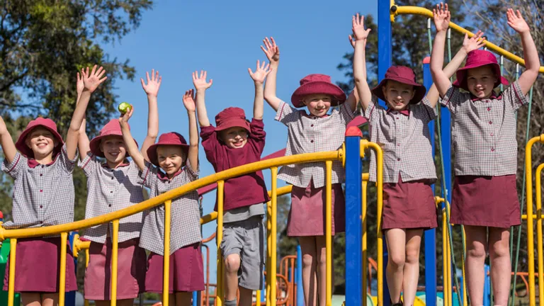 Group of children in maroon hats and checkered uniforms on a yellow-rail playground, arms raised.