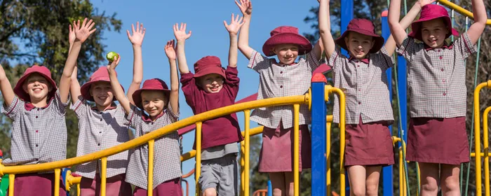 Group of children in maroon hats and checkered uniforms on a yellow-rail playground, arms raised.