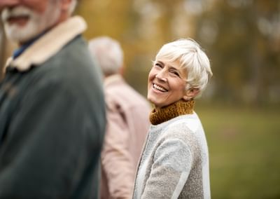 Smiling older person with short white hair in a grey-beige sweater, outdoors in a park.