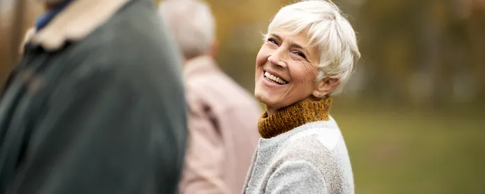 Smiling older person with short white hair in a grey-beige sweater, outdoors in a park.