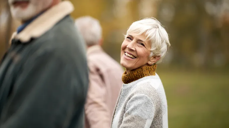 Smiling older person with short white hair in a grey-beige sweater, outdoors in a park.