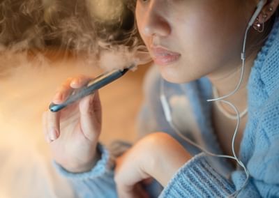 Close-up of a person in a blue hoodie vaping from a vape pen, wearing earphones.