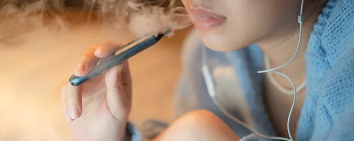 Close-up of a person in a blue hoodie vaping from a vape pen, wearing earphones.
