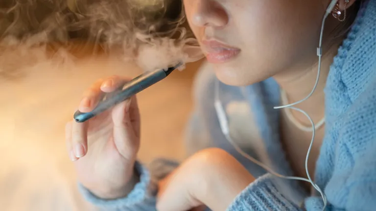 Close-up of a person in a blue hoodie vaping from a vape pen, wearing earphones.