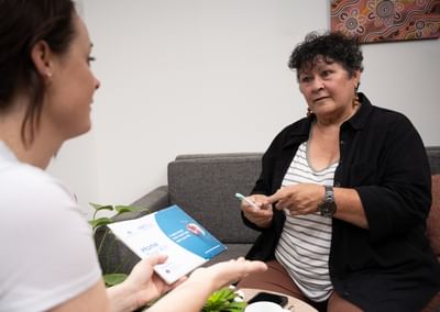 Two people on a grey sofa; one holds a blue and white brochure, the other points with a pen.