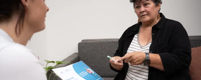 Two people on a grey sofa; one holds a blue and white brochure, the other points with a pen.