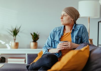 Person wearing headscarf and denim jacket sits on a sofa, holding a mug, with plants in living room.