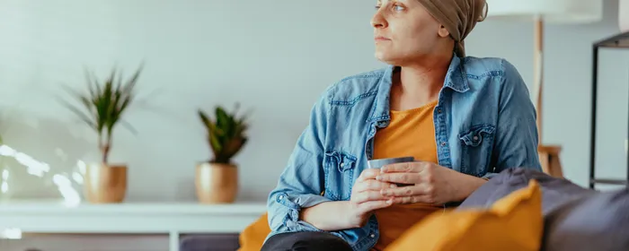 Person wearing headscarf and denim jacket sits on a sofa, holding a mug, with plants in living room.