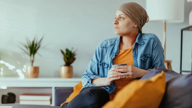 Person wearing headscarf and denim jacket sits on a sofa, holding a mug, with plants in living room.
