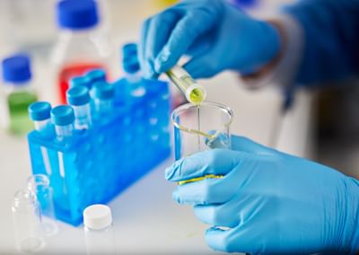 Gloved hands pour yellow liquid from a vial into a test tube in a lab with blue tube racks.