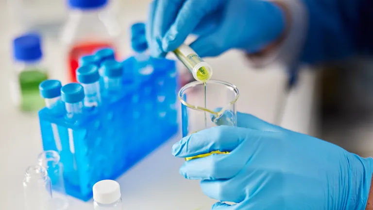 Gloved hands pour yellow liquid from a vial into a test tube in a lab with blue tube racks.