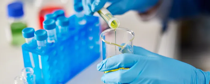 Gloved hands pour yellow liquid from a vial into a test tube in a lab with blue tube racks.