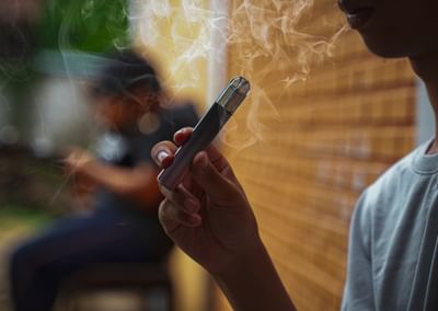 Person in foreground holding a vape pen with smoke; blurred figure and brick wall behind.