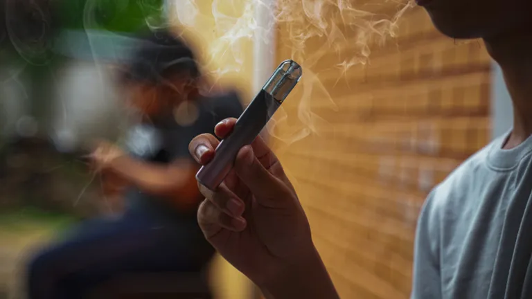 Person in foreground holding a vape pen with smoke; blurred figure and brick wall behind.
