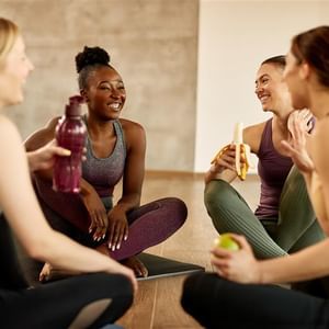Four people in athletic wear sit on yoga mats, smiling and chatting during a fitness break.