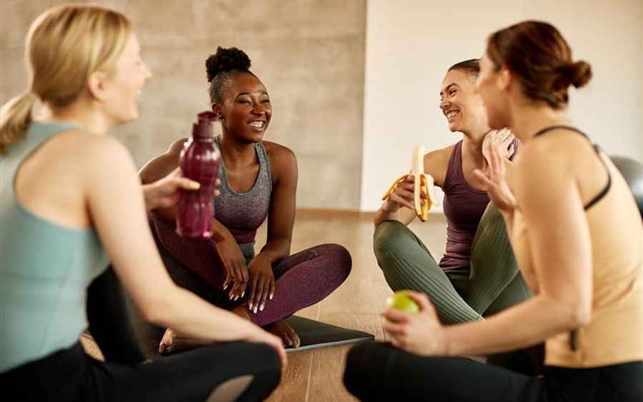 Four people in athletic wear sit on yoga mats, smiling and chatting during a fitness break.