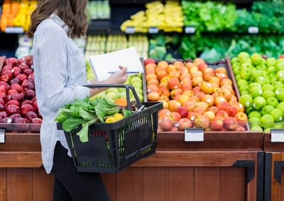 Person in a striped shirt with a black basket, taking notes while selecting fresh fruit and greens.