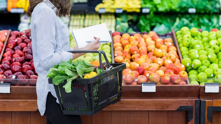 Person in a striped shirt with a black basket, taking notes while selecting fresh fruit and greens.