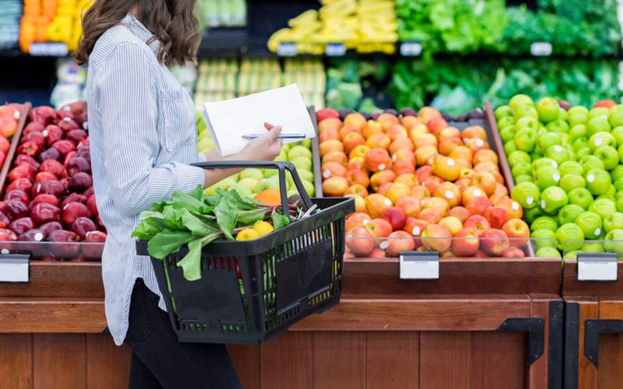 Person in a striped shirt with a black basket, taking notes while selecting fresh fruit and greens.