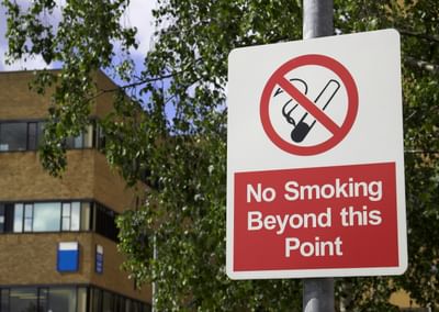 No smoking beyond this point sign on a post in front of a brick building and trees.