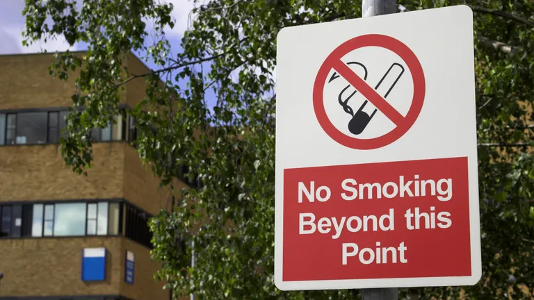 No smoking beyond this point sign on a post in front of a brick building and trees.