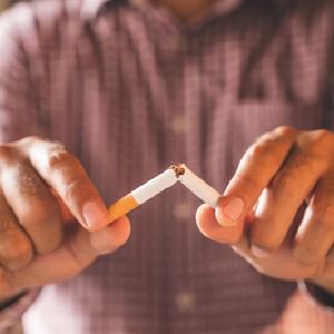 Person's hands breaking a cigarette in two, with a lit tip and ash, wearing a checkered shirt.