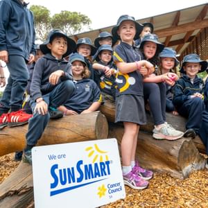 Group of schoolchildren in navy uniforms and bucket hats sit on logs beside a SunSmart sign.