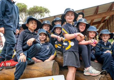 Group of schoolchildren in navy uniforms and bucket hats sit on logs beside a SunSmart sign.