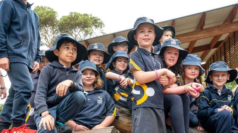 Group of schoolchildren in navy uniforms and bucket hats sit on logs beside a SunSmart sign.