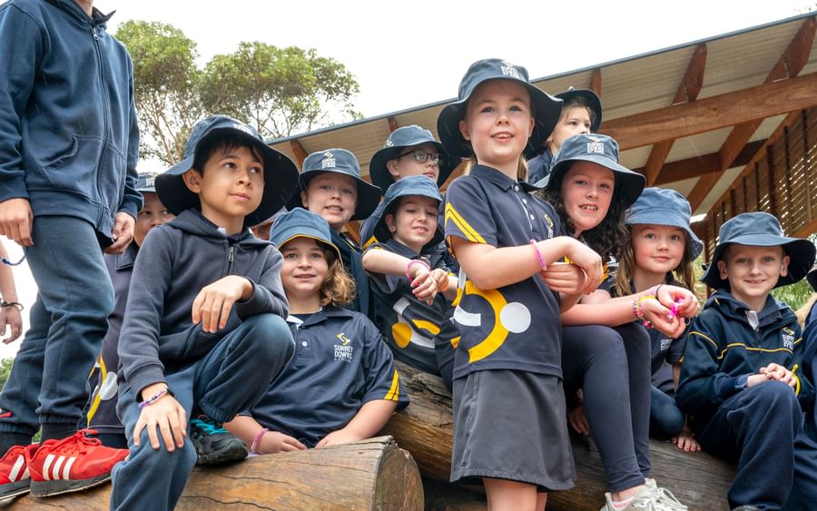 Group of schoolchildren in navy uniforms and bucket hats sit on logs beside a SunSmart sign.