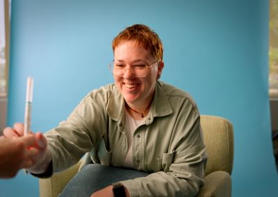 Smiling person with red hair and glasses shakes hands with another person beside a blue backdrop.