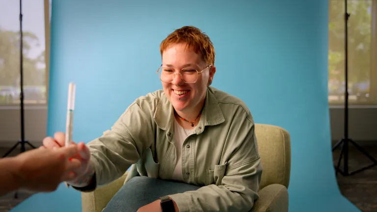 Smiling person with red hair and glasses shakes hands with another person beside a blue backdrop.
