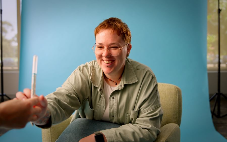 Smiling person with red hair and glasses shakes hands with another person beside a blue backdrop.