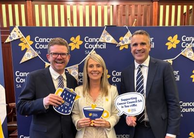 Three people in formal attire pose at a blue Cancer Council SA backdrop, holding props.