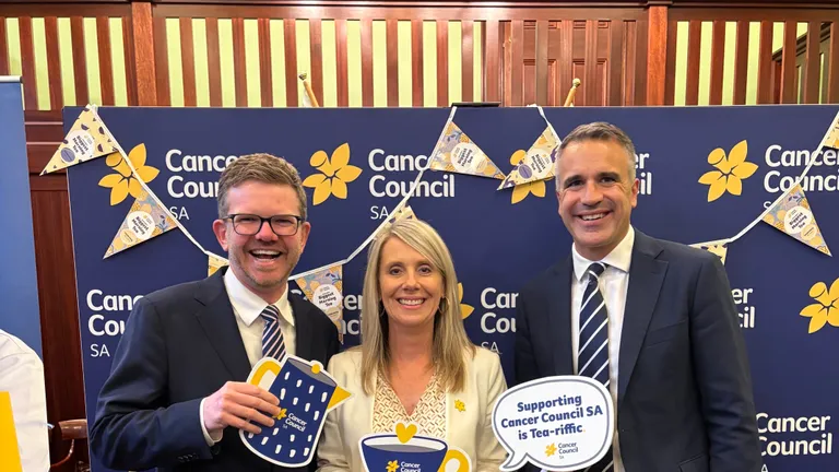 Three people in formal attire pose at a blue Cancer Council SA backdrop, holding props.