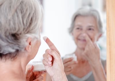 Older person looking into a mirror, applying cream to their nose while holding a jar.